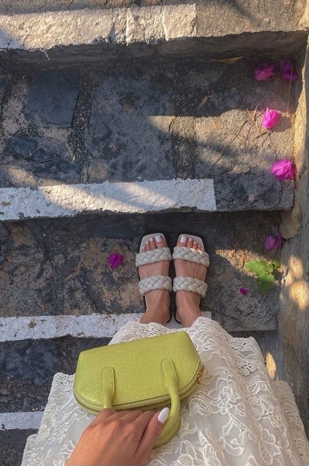 A first-person view looking down at feet in white braided sandals and a lime green handbag, with pink flower petals scattered on stone steps.