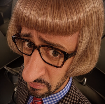 A close-up of a character with a brown bowl haircut, thick glasses, and a small mustache, wearing a checkered shirt and tie.