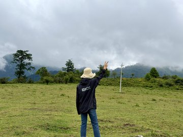 A person wearing a dark "OKX" jacket and a wide-brimmed hat stands in a grassy meadow, reaching out toward misty mountains under an overcast sky, embodying the post's invitation to enjoy the outdoors in a relaxed, on-the-go market vibe.