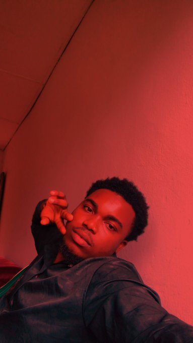 A low-angle selfie of a young man wearing a dark hoodie, bathed in intense red lighting against a plain wall, posing with his hand reaching toward the camera.