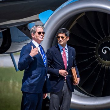 Two men in suits and sunglasses standing in front of a large aircraft jet engine, one giving a thumbs-up.