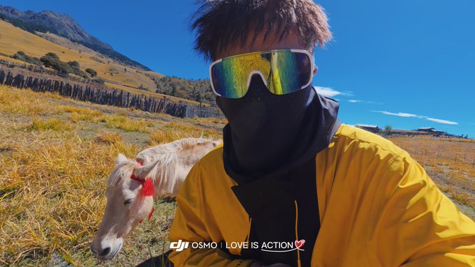 A selfie taken outdoors showing a person wearing a yellow jacket, black face covering, and reflective sunglasses, posing next to a small white pony in a grassy mountainous landscape, with a 'DJI OSMO | LOVE IS ACTION' watermark.