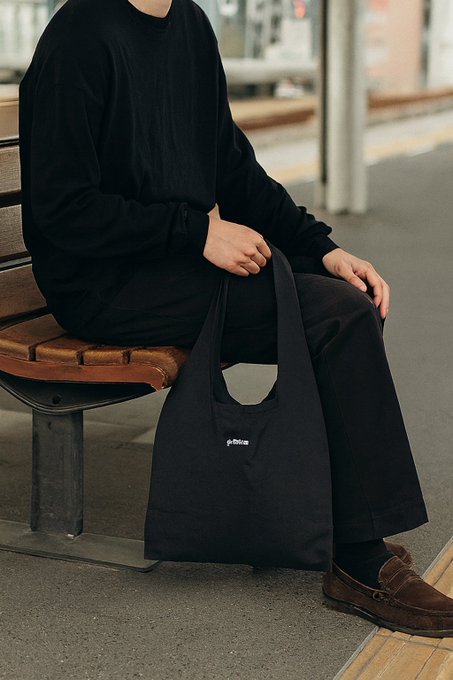 A person in all-black clothing sitting on a wooden bench holding a black tote bag with a small "gd frogs" label, wearing brown suede loafers.