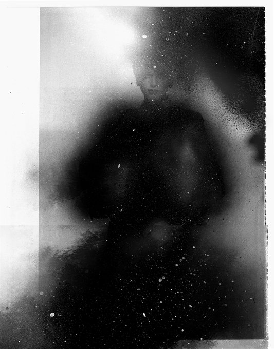 An artistic black-and-white portrait features a person obscured by blurry, smoky effects and dark clothing against a dark background, with only faint facial features visible.