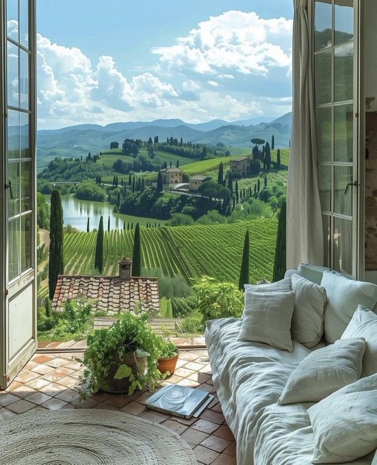 A view through an open French window onto a Tuscan-style landscape with rolling green hills, vineyards, cypress trees, and a lake, with a daybed covered in white pillows in the foreground.