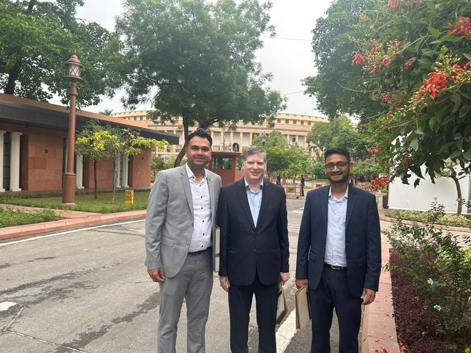 Photo of three men in business attire posing together outdoors in front of a large official-looking building, likely in India based on the architecture and signage.