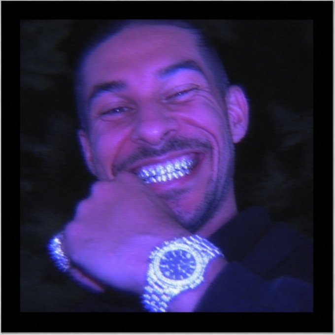 A close-up, purple-lit portrait of a smiling man showing off diamond-encrusted grills and a large, diamond-covered watch.
