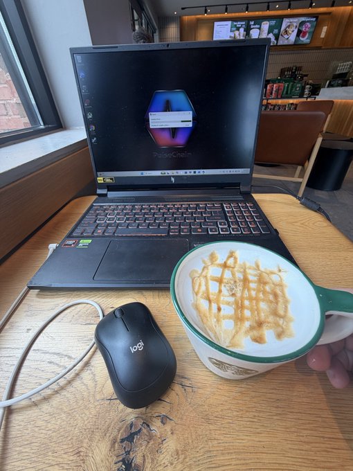 A wooden table at a café setting up a laptop displaying a Polygon logo, accompanied by a Logitech mouse and a hand holding a bowl topped with a waffle.