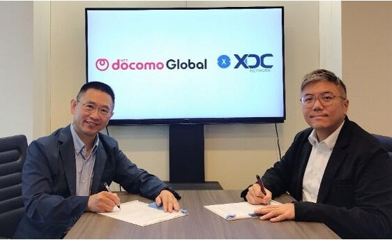 Two men in business attire sit at a conference table signing partnership documents, with a screen behind them displaying the NTT docomo Global and XDC Network logos, symbolizing XDC Network's new masternode validator partnership with NTT DOCOMO in Japan.