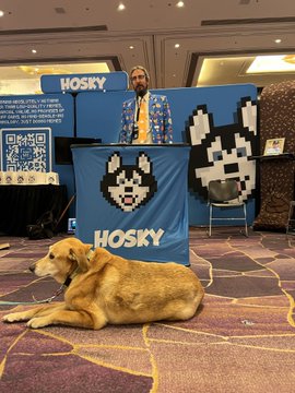 A man standing at a 'HOSKY' booth with a husky dog mascot logo, accompanied by a large golden retriever dog lying on the carpeted floor in front.