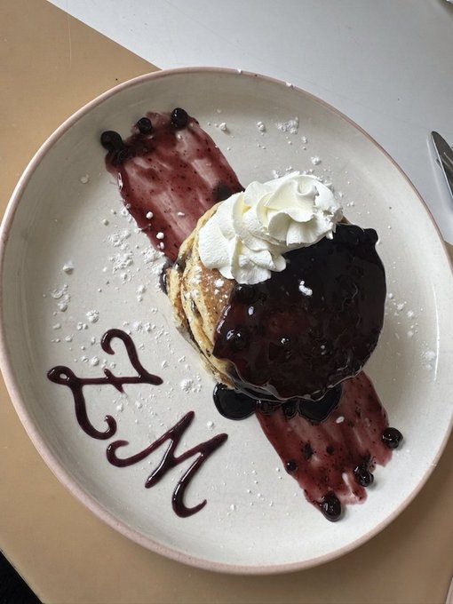 The image features a white plate with pancakes topped with blueberries, syrup, and whipped cream. The plate has the words "ZM" written on it in cursive font. The background of the image is a light beige color, which contrasts nicely with the white plate holding the delicious breakfast dish.