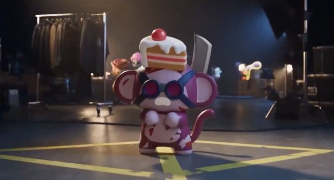 A small, colorful toy figure with a cake-like head and large round eyes stands on a dark surface marked with yellow tape, set in a dimly lit industrial space.