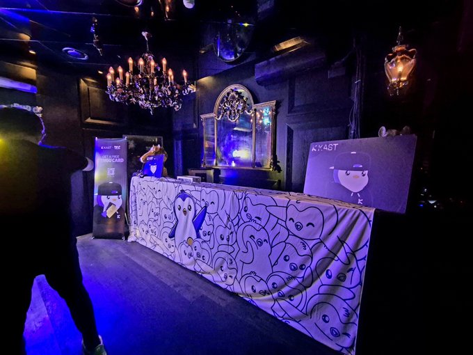 A dimly lit venue featuring an ornate chandelier and gothic-style mirror sets a moody, party-like atmosphere, with a check-in counter branded with "KAST" signage and Pudgy Penguin artwork on the front, alongside a "Pudgy Penguin Card Party" banner.