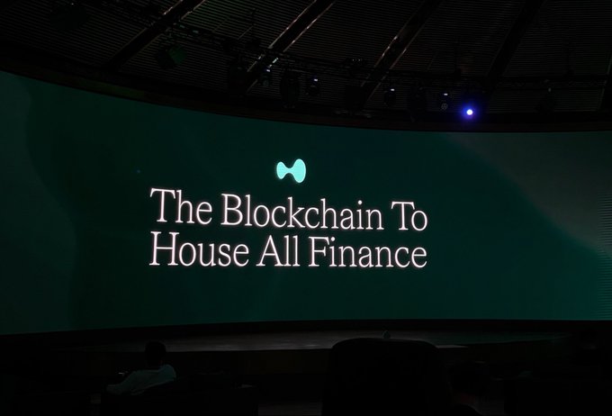A presentation slide displayed on a large screen reading "The Blockchain To House All Finance" with a green logo above it.