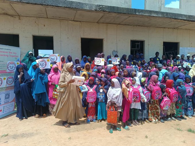 A large group of girls and women in colorful hijabs and backpacks gathered outside a building, participating in a "Scorefor the Joy of Learning Together" educational program, holding smiley face signs.