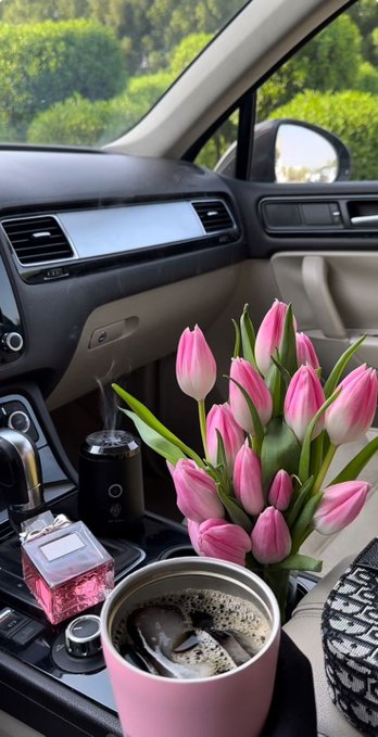 Interior view of a car featuring a bouquet of pink tulips in the passenger seat alongside a pink cup of coffee, a tumbler, and a pink perfume bottle, with greenery visible through the windshield.