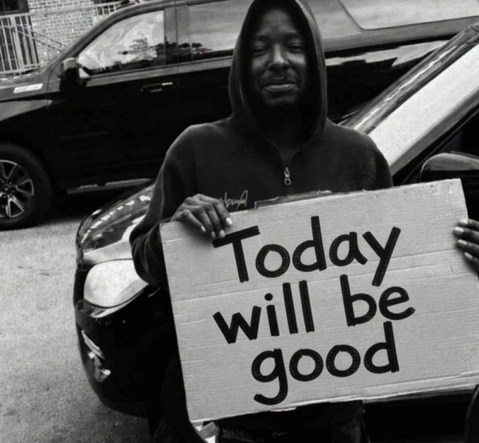 A black and white photo of a man in a hooded sweatshirt standing in front of a black car, holding a cardboard sign that reads 'Today will be good'.