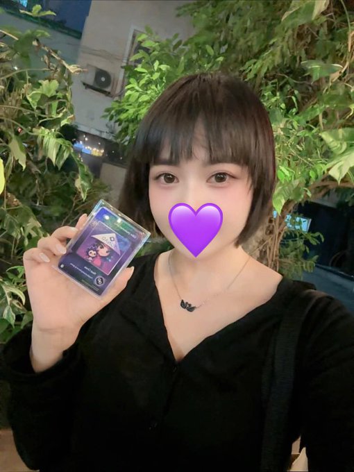 A young woman with short dark hair and a black top holds up a Pokémon trading card, with a purple heart emoji covering her mouth.