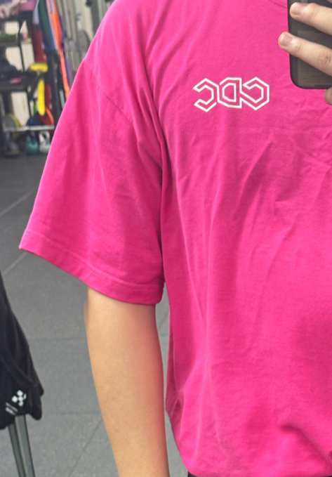 A person takes a mirror selfie wearing an oversized bright pink t-shirt featuring a stylized 'SOS' logo on the chest.