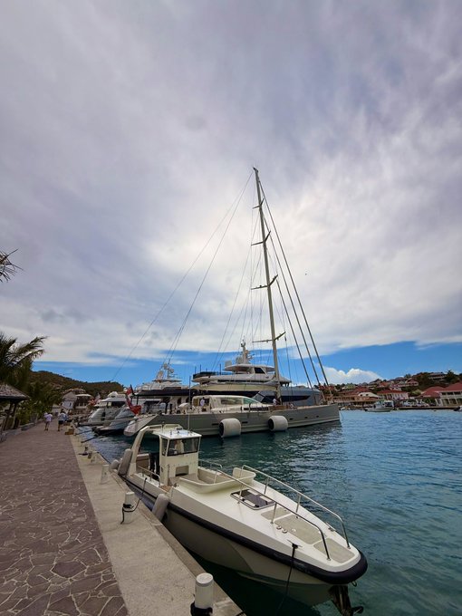The image shows a marina with several boats docked at the pier, including a small white boat near the center of the frame. The sky above appears cloudy, creating an overcast atmosphere. In the background, there are two larger boats visible - one on the left side and another towards the right edge of the image.