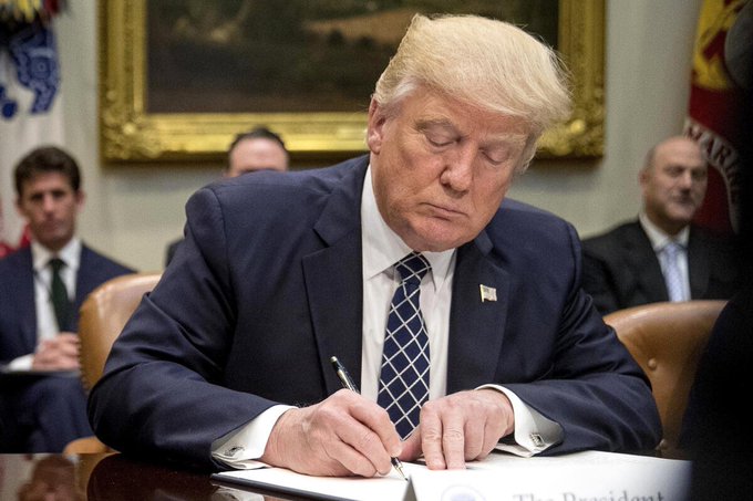 Donald Trump, identified by the "The President" nameplate visible on the desk, is shown signing a document in the Oval Office, illustrating his enactment of legislation imposing up to 100% tariffs on nations purchasing Russian oil.