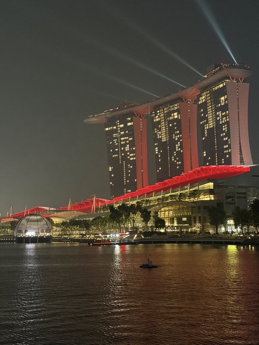 A nighttime photograph of the Marina Bay Sands hotel in Singapore, illuminated in red and gold lights with beams shining upward, reflected in the water below.