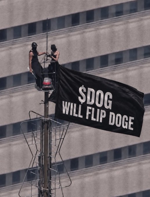 Two masked individuals stand on a platform atop a communication tower, unfurling a large black banner that reads "$DOG WILL FLIP DOGE," directly showcasing the bold cryptocurrency prediction in a dramatic urban display.