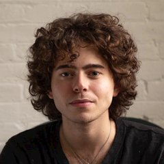 A young man with curly brown hair wearing a dark shirt and a thin necklace poses against a white brick wall in this portrait. The image appears to accompany an article about Polymarket CEO Shayne Coplan's comments comparing crypto speculation to "irrational exuberance and hot potato," though no identifying text appears in the photo itself.