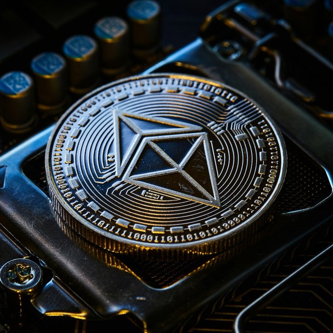 A physical silver Ethereum (ETH) coin rests on a computer circuit board, with the binary code ring around the coin and the geometric Ethereum logo directly tying the visual to the cryptocurrency's digital infrastructure. This imagery complements the news of Bitmine expanding its Ethereum holdings to over 5.9 million ETH, symbolizing the growing institutional accumulation of the asset.
