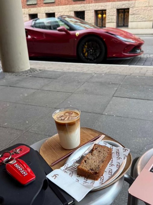 An outdoor cafe scene featuring an iced coffee and a slice of cake on a plate, with a red Ferrari sports car parked on the street in the background and a Ferrari key fob visible on the table.