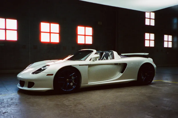 A white Porsche Carrera GT convertible parked in a dark garage illuminated by red and white square windows on the back wall.