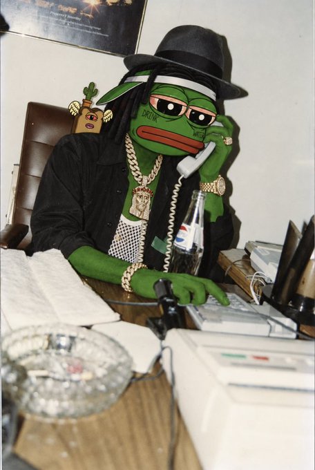 A person in a Pepe the Frog mask, black hat, and jacket with gold chains, sitting at a desk holding a telephone receiver, with a Pepsi bottle and an ashtray visible.
