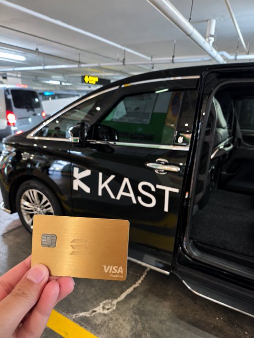 A hand holds a gold metal Visa card branded with 'KAST' in the foreground, with a matching black luxury vehicle with 'KAST' lettering visible inside a parking garage.
