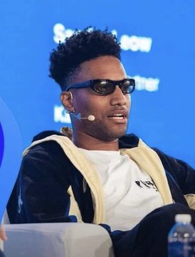 A man with curly hair, wearing sunglasses and a headset microphone, is speaking at what appears to be a conference panel, with partial text visible in the background suggesting a partnership-related discussion. The image relates to the post about ANSEM's commentary on PumpFun's potential move toward creating a stablecoin rather than launching an L1 blockchain.