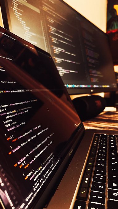A close-up, angled view of a laptop screen displaying lines of code, with an external monitor also showing code in the blurred background.