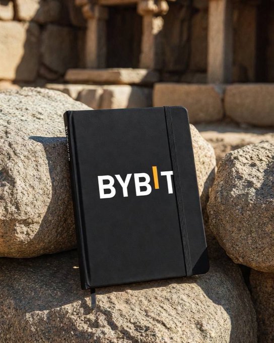 A black notebook branded with the 'BYBIT' logo rests against rough gray stones in what appears to be ancient ruins bathed in warm sunlight.