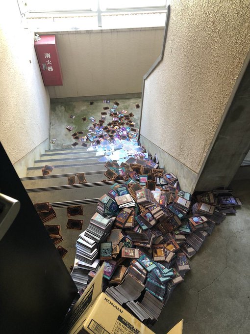 A stairwell filled with a massive pile of trading cards, predominantly in protective sleeves, scattered down the steps with a red fire equipment box on the wall.