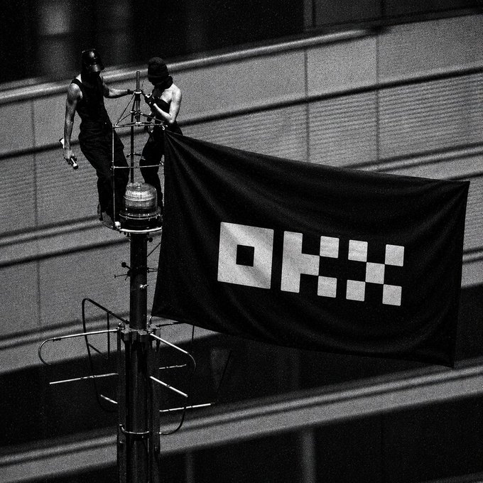 Two workers atop a tall pole attach a large flag reading "OKX" (referencing the cryptocurrency exchange), visually echoing the post "GM 高处见见未来" (Good Morning, see the future from the heights). The high vantage point of the installation symbolizes forward-looking ambition in the crypto industry.