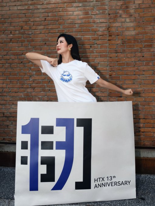 A woman in a white "INUBAO" t-shirt poses behind a large banner for the "HTX 13th ANNIVERSARY" celebration. The banner features large blue and black Chinese characters and the text "HTX 13th ANNIVERSARY", connecting to the post's announcement of an event at 8 PM on @HTX_Live.