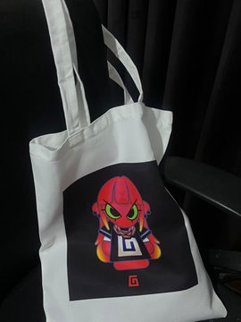 A white canvas tote bag features a printed graphic of a red cartoon character on a black square background, with a small 'G' logo in the corner.