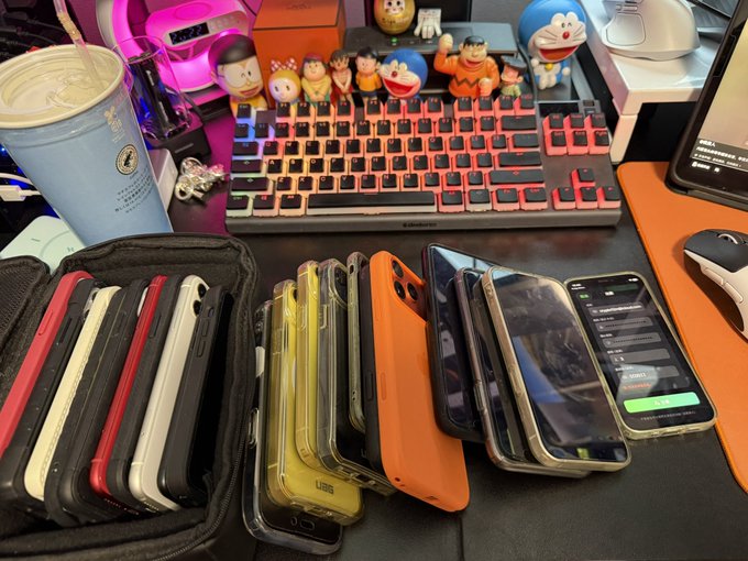 A desk scene showing multiple smartphones stored in a black case alongside additional phones with colorful cases (yellow, orange, black) laid out next to a mechanical keyboard and a coffee cup; the accompanying post references iPhone opportunities, tying into the image as the poster displays various iPhones (including ones running messaging apps) likely being prepared for sale or trade.