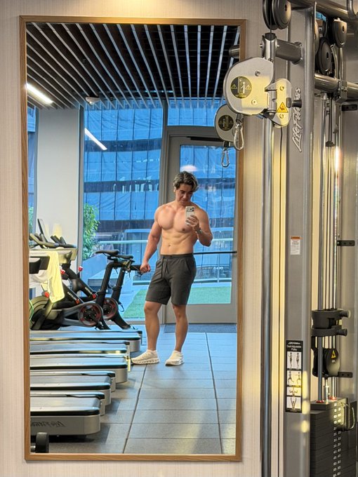 A mirror photo of a muscular shirtless man in dark shorts taking a selfie in a gym, with exercise equipment and treadmills visible around him.