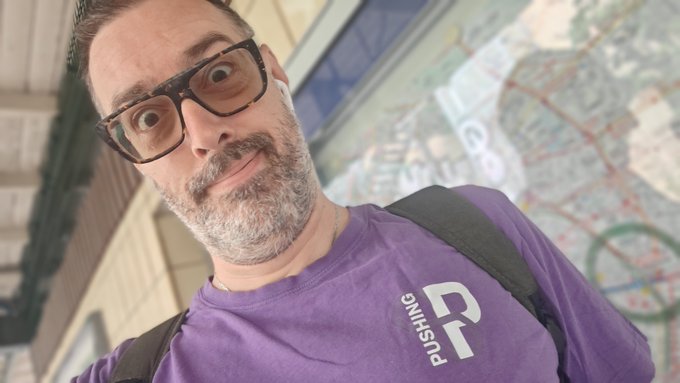 A selfie of a man with a beard and glasses wearing a purple shirt that says 'PLUSHKING' with a 'P' logo, standing in front of a map board.
