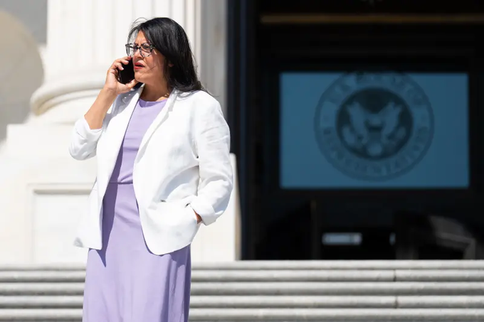 A woman in a white blazer and lavender dress talks on her phone while standing on the steps of a building with the United States House of Representatives seal visible on the door behind her.