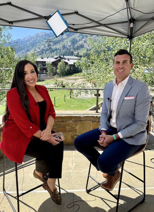 A woman in a red blazer and a man in a blue suit sit on stools under a white tent for an outdoor interview, with a scenic mountain valley visible behind them. A small digital display in the upper left shows partially legible text that appears to read "OST" (likely part of a larger word). The setup matches the post's description of an interview with CNBC's Talia Kaplan at the SALT Conference Wyoming Blockchain Symposium.