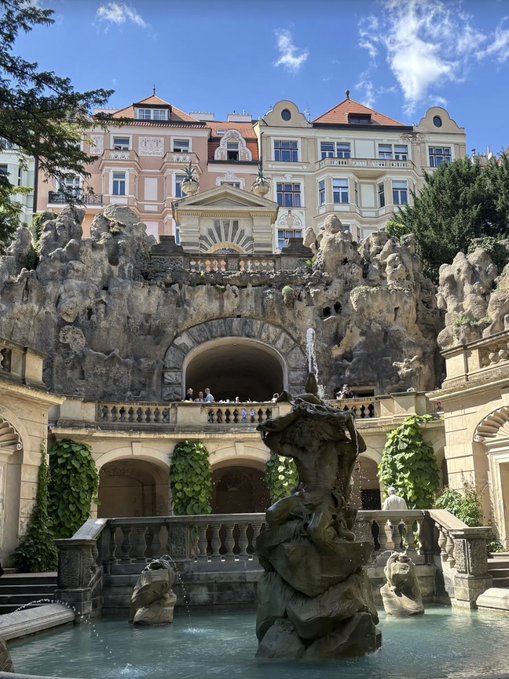 The image shows an outdoor courtyard with a large stone fountain surrounded by several people sitting around it, enjoying their time. The fountain has a statue of a mermaid perched atop one of its rocks. In the background, there are buildings that add depth to the scene and provide context for the location. A post on social media is visible in the image, likely sharing details about the beautiful outdoor space or inviting viewers to visit the site as well.