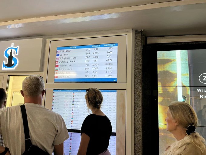 Three people stand before digital screens at what appears to be a currency exchange booth, with the top display showing USD to PLN rates at 4.375 and EUR to PLN at 4.665, alongside a logo featuring "SK" letters. The favorable exchange rates displayed support the sentiment expressed in the post about the currency conversion being a good deal.