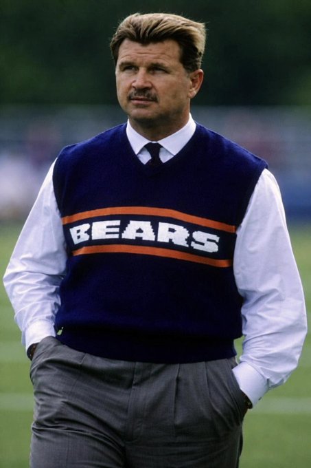 A man with a mustache stands on a football field wearing a blue Chicago Bears sweater vest over a white dress shirt and tie. The "BEARS" text on his vest, combined with the tribute message, suggests this image honors the legendary NFL coach known as "Da Coach."