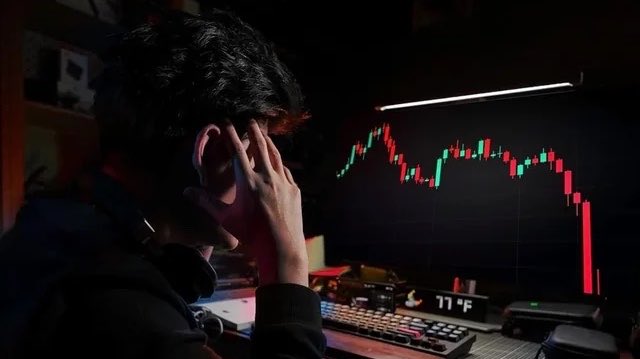 A person sits hunched in front of a computer monitor displaying a sharply falling candlestick chart in red and green, reflecting the massive liquidation of 102,332 crypto traders in the past 24 hours.