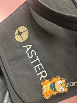 A black leather bag with "ASTER" printed in gold lettering sits next to a cute orange cat keychain, likely the giveaway mentioned from Aster's private event.